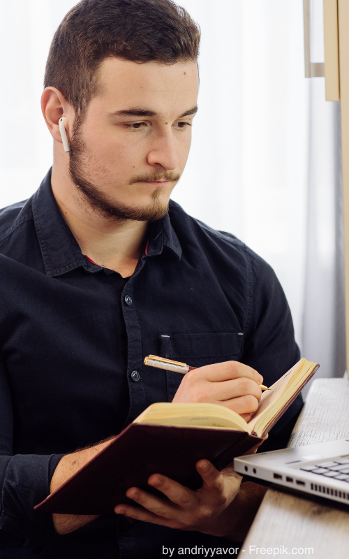 Ein junger Mann mit Kopfhörern vor dem Laptop macht sich Notizen.