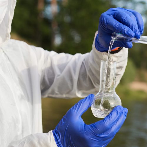 Water Purity Test. Hand in protective gloves holding a chemical flask or test tube with water. Lake in the background.